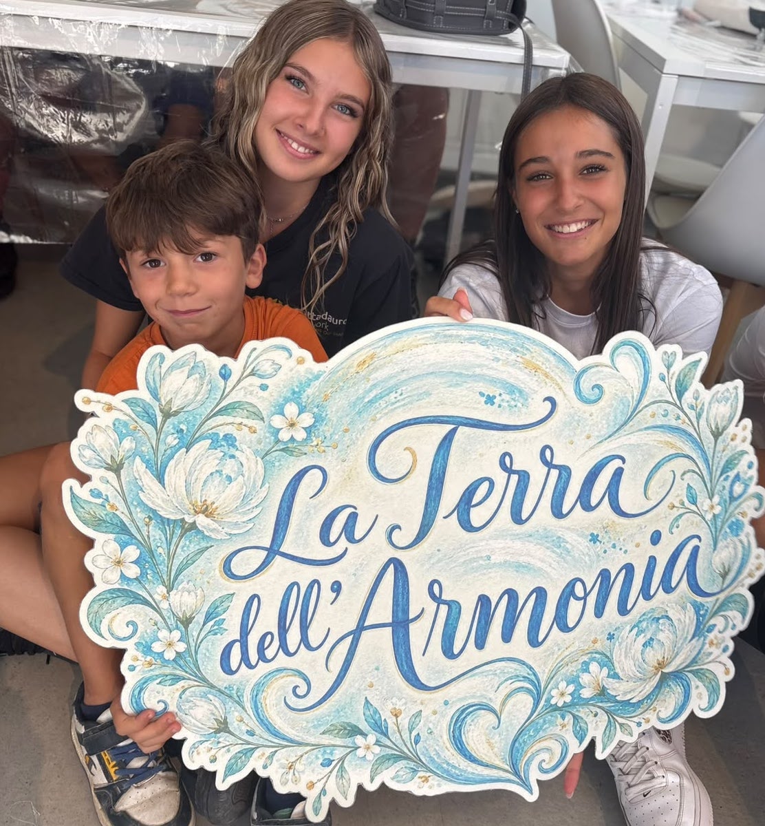 Tre ragazzi sorridono mostrando l'insegna dipinta La Terra dell'Armonia